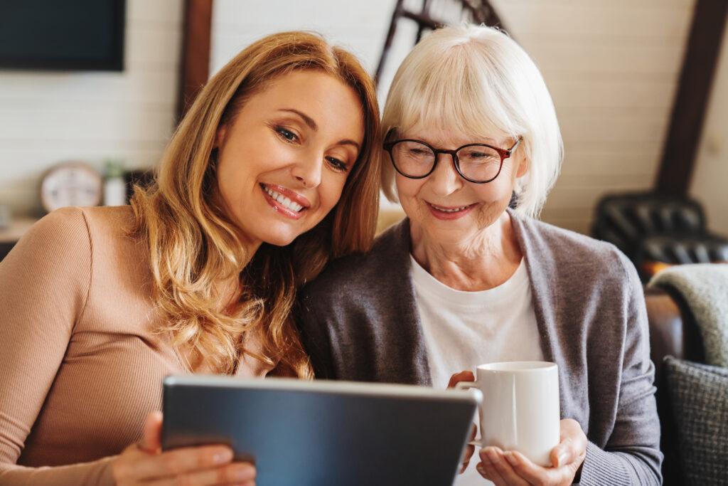 Fröhliche Seniorin mit einer Tasse Tee und ihrer Tochter, die zu Hause ein Tablet benutzt